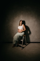 A pregnant woman is sitting on a small ladder, holding her belly with both hands. She is smiling and wearing a white top and a light green skirt. The background consists of a plain, textured gray wall. The lighting casts a soft shadow behind her, creating an intimate and warm atmosphere.