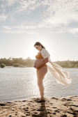 A pregnant woman stands by a serene body of water, silhouetted against the sun. She holds her belly gently, wearing a light-colored bikini and a sheer cover-up that drapes behind her in the breeze. The sand and the calm water suggest a peaceful outdoor setting, with a soft, cloudy sky overhead.