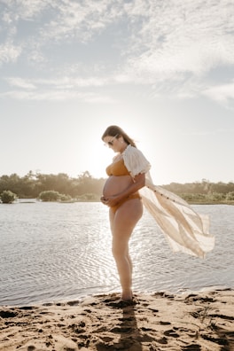 A pregnant woman stands by a serene body of water, silhouetted against the sun. She holds her belly gently, wearing a light-colored bikini and a sheer cover-up that drapes behind her in the breeze. The sand and the calm water suggest a peaceful outdoor setting, with a soft, cloudy sky overhead.
