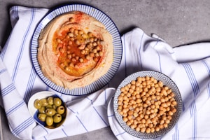 A flatlay of a table setting featuring various food items, primarily featuring a bowl of creamy hummus garnished with chickpeas and spices, a bowl of chickpeas, and a small bowl of green olives. The items are set on a white and blue striped cloth with a concrete surface background.