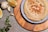 A bowl of creamy hummus topped with chickpeas and spices is placed on a blue and white plate, which rests on a wooden board. Surrounding the bowl are lemon wedges, cloves of garlic, and slices of green pepper, with scattered chili flakes on a textured gray surface.