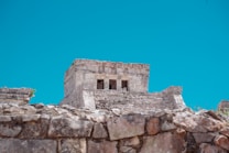 Ancient stone ruins with a tower-like structure and three windows on top are set against a clear blue sky. The architecture features weathered stones and sharp lines, indicating advanced construction techniques.