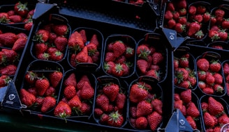 A collection of dark plastic trays filled with ripe, red strawberries arranged neatly. The strawberries have a vibrant red color, with small green leafy tops, and are displayed in a symmetric pattern. The setting appears to be in a market or a store, with each tray being labeled and organized for sale.