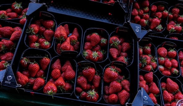 A collection of dark plastic trays filled with ripe, red strawberries arranged neatly. The strawberries have a vibrant red color, with small green leafy tops, and are displayed in a symmetric pattern. The setting appears to be in a market or a store, with each tray being labeled and organized for sale.