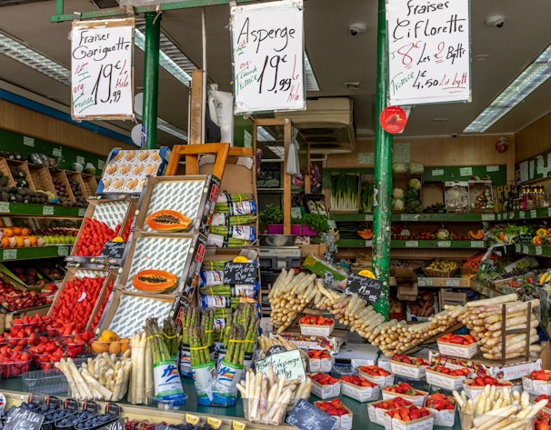 A vibrant market stall filled with a variety of fresh produce including asparagus, strawberries, and other fruits and vegetables. The setup includes wooden crates and baskets showcasing the colorful array of items. Price tags are prominently displayed in handwritten style on signs above the produce.