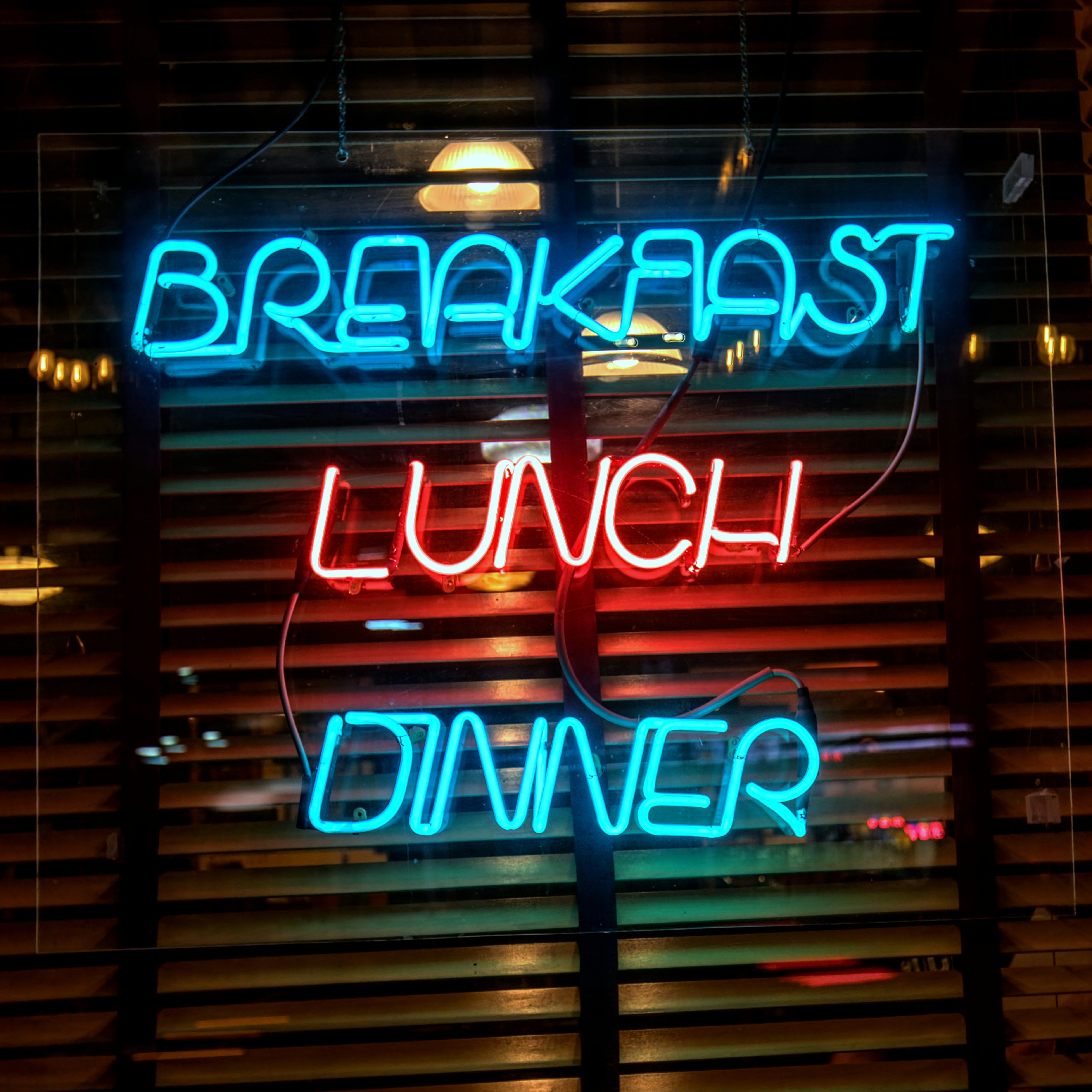An image of a window with blinds at night, with a neon sign that says “Breakfast Lunch Dinner”