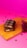 A partially eaten chocolate bar rests on a wooden board, revealing a gooey caramel and nougat filling. The background is a vibrant pink, scattered with chocolate chips and small clusters of dark pink flowers.