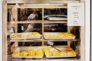 A variety of baked goods are displayed in metal trays within a glass display case. The baked items appear to be buns with a golden-brown crust. A sign with pricing information is attached to the right side of the display. The reflection of a person taking a photograph is visible in the glass.