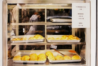 A variety of baked goods are displayed in metal trays within a glass display case. The baked items appear to be buns with a golden-brown crust. A sign with pricing information is attached to the right side of the display. The reflection of a person taking a photograph is visible in the glass.