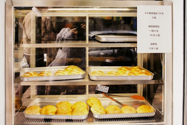 A variety of baked goods are displayed in metal trays within a glass display case. The baked items appear to be buns with a golden-brown crust. A sign with pricing information is attached to the right side of the display. The reflection of a person taking a photograph is visible in the glass.