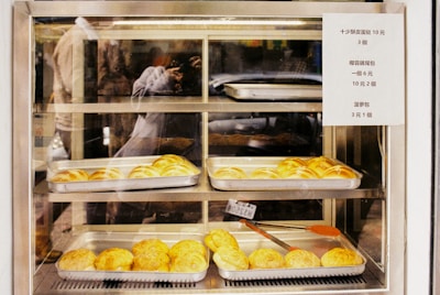 A variety of baked goods are displayed in metal trays within a glass display case. The baked items appear to be buns with a golden-brown crust. A sign with pricing information is attached to the right side of the display. The reflection of a person taking a photograph is visible in the glass.
