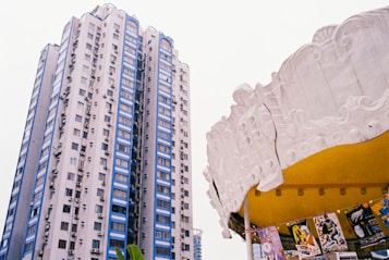 A tall residential building with a modern design features blue accents and numerous windows. In the foreground, part of a carousel with ornate white and yellow detailing displays colorful posters.