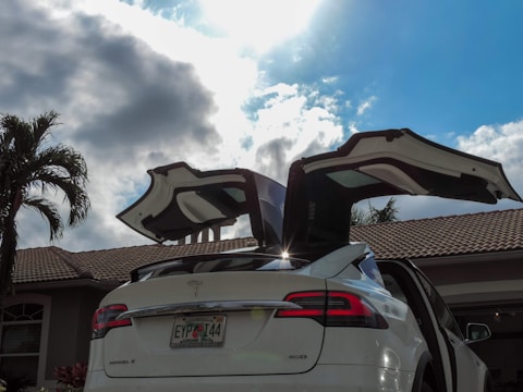A white car is parked in front of a house with open falcon-wing doors. The vehicle is situated under a partly cloudy sky with sunlight shining through. There is a palm tree to the left and a tiled roof of the house is visible in the background.