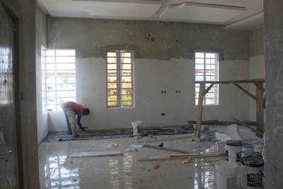 A room under renovation with unfinished walls and windows. A person is working on the floor, surrounded by construction materials including paint buckets, wooden planks, and plastic sheets. Sunlight streams in through the large windows, casting shadows on the partially plastered walls.
