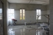 A room under renovation with unfinished walls and windows. A person is working on the floor, surrounded by construction materials including paint buckets, wooden planks, and plastic sheets. Sunlight streams in through the large windows, casting shadows on the partially plastered walls.