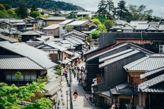 An aerial view of a traditional Japanese street lined with wooden houses, featuring tiled roofs and surrounded by lush greenery and distant hills. People walk along the narrow pathways between the houses, creating a bustling yet serene atmosphere. The architecture is classic, with an emphasis on natural materials and harmony with the environment.