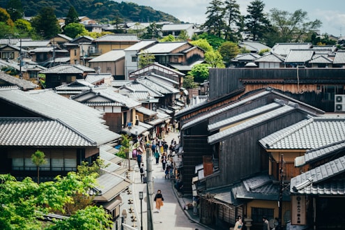 An aerial view of a traditional Japanese street lined with wooden houses, featuring tiled roofs and surrounded by lush greenery and distant hills. People walk along the narrow pathways between the houses, creating a bustling yet serene atmosphere. The architecture is classic, with an emphasis on natural materials and harmony with the environment.