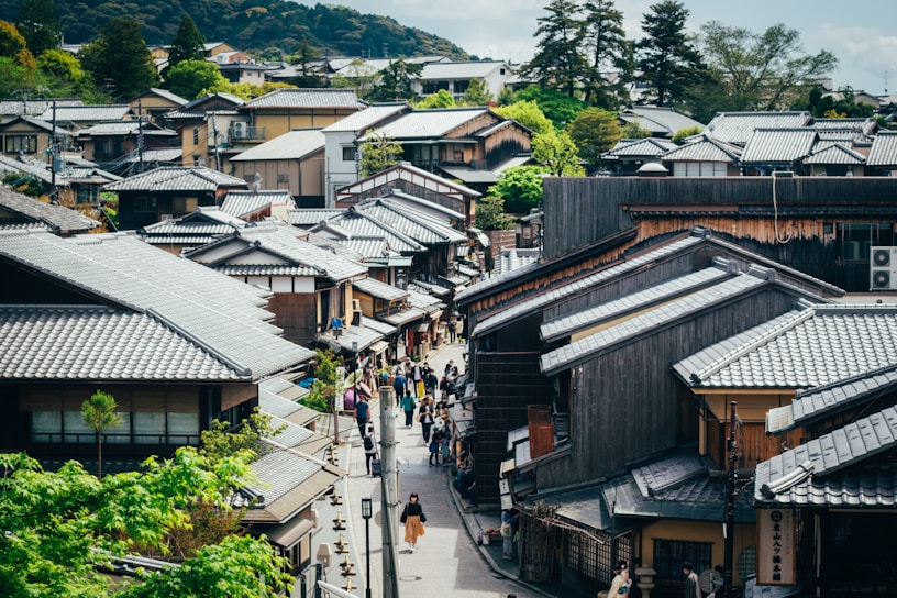 An aerial view of a traditional Japanese street lined with wooden houses, featuring tiled roofs and surrounded by lush greenery and distant hills. People walk along the narrow pathways between the houses, creating a bustling yet serene atmosphere. The architecture is classic, with an emphasis on natural materials and harmony with the environment.