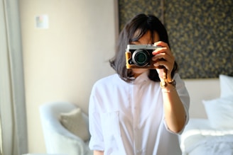 A person with medium-length dark hair is holding a camera, obscuring their face. They are wearing a white shirt and standing in a bright room with a patterned wall in the background. Soft natural light is highlighting their features and casting shadows.