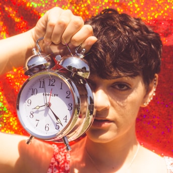 A person with short, dark hair holds a large, silver alarm clock up to their face. The background is a vibrant, sparkling red, creating a striking contrast with the clock's metallic finish. The person is dressed in a red-patterned outfit, complementing the background color.