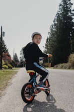 A young child wearing a cap is riding a small bicycle on a quiet road lined with tall evergreen trees. The child is turning to look back, while the sun casts soft shadows on the ground. The bicycle features bright orange wheels and blue accents. Surrounding the road, there is green grass and distant utility poles.