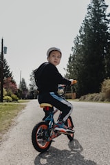 A young child wearing a cap is riding a small bicycle on a quiet road lined with tall evergreen trees. The child is turning to look back, while the sun casts soft shadows on the ground. The bicycle features bright orange wheels and blue accents. Surrounding the road, there is green grass and distant utility poles.