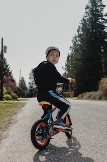 A young child wearing a cap is riding a small bicycle on a quiet road lined with tall evergreen trees. The child is turning to look back, while the sun casts soft shadows on the ground. The bicycle features bright orange wheels and blue accents. Surrounding the road, there is green grass and distant utility poles.