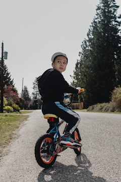 A young child wearing a cap is riding a small bicycle on a quiet road lined with tall evergreen trees. The child is turning to look back, while the sun casts soft shadows on the ground. The bicycle features bright orange wheels and blue accents. Surrounding the road, there is green grass and distant utility poles.