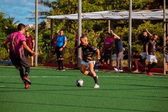 A youth soccer match is taking place on an outdoor field. A player in a black jersey is in motion, dribbling the ball, while a player in a pink jersey approaches from the left. Several people are watching from the sidelines, some in casual clothing and others wearing jerseys. The field is surrounded by a chain-link fence, with greenery visible in the background. A referee in a blue shirt stands near the sideline, observing the game.