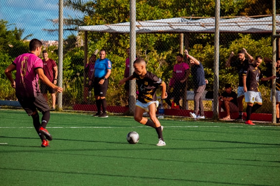 A youth soccer match is taking place on an outdoor field. A player in a black jersey is in motion, dribbling the ball, while a player in a pink jersey approaches from the left. Several people are watching from the sidelines, some in casual clothing and others wearing jerseys. The field is surrounded by a chain-link fence, with greenery visible in the background. A referee in a blue shirt stands near the sideline, observing the game.