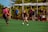 A youth soccer match is taking place on an outdoor field. A player in a black jersey is in motion, dribbling the ball, while a player in a pink jersey approaches from the left. Several people are watching from the sidelines, some in casual clothing and others wearing jerseys. The field is surrounded by a chain-link fence, with greenery visible in the background. A referee in a blue shirt stands near the sideline, observing the game.