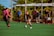A youth soccer match is taking place on an outdoor field. A player in a black jersey is in motion, dribbling the ball, while a player in a pink jersey approaches from the left. Several people are watching from the sidelines, some in casual clothing and others wearing jerseys. The field is surrounded by a chain-link fence, with greenery visible in the background. A referee in a blue shirt stands near the sideline, observing the game.