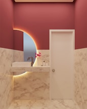 A modern bathroom with marble-patterned walls and floor, featuring a white sink with a sleek faucet. A circular mirror with an illuminated backlit design is present, and a small arrangement of pink artificial flowers is placed on the countertop along with a soap dispenser and a bottle.