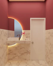 A modern bathroom with marble-patterned walls and floor, featuring a white sink with a sleek faucet. A circular mirror with an illuminated backlit design is present, and a small arrangement of pink artificial flowers is placed on the countertop along with a soap dispenser and a bottle.