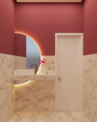 A modern bathroom with marble-patterned walls and floor, featuring a white sink with a sleek faucet. A circular mirror with an illuminated backlit design is present, and a small arrangement of pink artificial flowers is placed on the countertop along with a soap dispenser and a bottle.