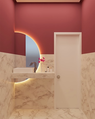A modern bathroom with marble-patterned walls and floor, featuring a white sink with a sleek faucet. A circular mirror with an illuminated backlit design is present, and a small arrangement of pink artificial flowers is placed on the countertop along with a soap dispenser and a bottle.