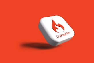 A white, square button with rounded edges featuring a red flame logo and the word 'CodeIgniter' printed beneath it. The button casts a shadow on an orange background, creating a clean and modern design.