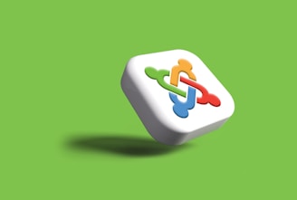 A three-dimensional square with rounded edges featuring an interlocking design in red, green, blue, and orange on its front, set against a vibrant green background. The shadow underneath suggests the object is floating.