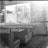 A black and white photograph of a retro diner interior. Booth seating with a table and napkin dispenser is visible in the foreground. Vintage posters and advertisements cover the walls, alongside large windows letting in bright natural light, creating a nostalgic and timeless atmosphere.