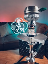 A modern hookah with a decorative bowl and hose is placed on a table. In the background, there is a couch and a neon light shaped like a hookah, emitting a colorful glow. The room has a relaxed and cozy atmosphere.