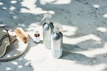 A set of cleaning products is arranged on a light-colored marble surface. The items include two metal spray bottles, a small glass bottle with a liquid, a natural bristle brush, and a cloth with dark stripes. Sunlight filters through leaves, casting dappled shadows on the surface.