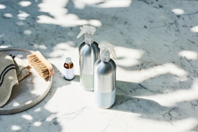 A set of cleaning products is arranged on a light-colored marble surface. The items include two metal spray bottles, a small glass bottle with a liquid, a natural bristle brush, and a cloth with dark stripes. Sunlight filters through leaves, casting dappled shadows on the surface.