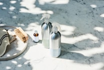 A set of cleaning products is arranged on a light-colored marble surface. The items include two metal spray bottles, a small glass bottle with a liquid, a natural bristle brush, and a cloth with dark stripes. Sunlight filters through leaves, casting dappled shadows on the surface.