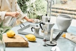 A person is cleaning a countertop with a cloth while holding a spray bottle. On the counter, there are two metal stovetop coffee makers, a white ceramic vase, a wooden cutting board with two lemons, and various kitchen items, including a candle and small bottle.