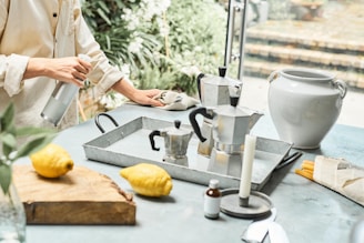 A person is cleaning a countertop with a cloth while holding a spray bottle. On the counter, there are two metal stovetop coffee makers, a white ceramic vase, a wooden cutting board with two lemons, and various kitchen items, including a candle and small bottle.