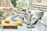A person is cleaning a countertop with a cloth while holding a spray bottle. On the counter, there are two metal stovetop coffee makers, a white ceramic vase, a wooden cutting board with two lemons, and various kitchen items, including a candle and small bottle.