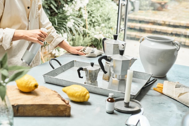A person is cleaning a countertop with a cloth while holding a spray bottle. On the counter, there are two metal stovetop coffee makers, a white ceramic vase, a wooden cutting board with two lemons, and various kitchen items, including a candle and small bottle.