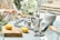 A person is cleaning a countertop with a cloth while holding a spray bottle. On the counter, there are two metal stovetop coffee makers, a white ceramic vase, a wooden cutting board with two lemons, and various kitchen items, including a candle and small bottle.