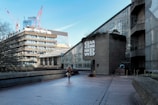 A modern urban setting features a large concrete building with Brutalist architecture, including a distinctive geometric design. A person is walking along a paved walkway. In the background, several cranes are visible, indicating construction activity in the area. The sky is clear and blue, and the atmosphere feels quiet and spacious.