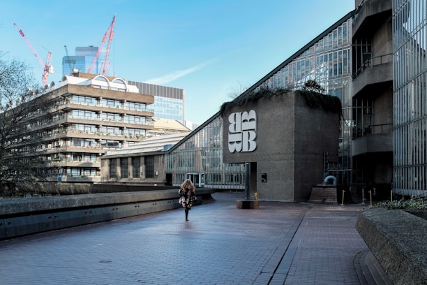 A modern urban setting features a large concrete building with Brutalist architecture, including a distinctive geometric design. A person is walking along a paved walkway. In the background, several cranes are visible, indicating construction activity in the area. The sky is clear and blue, and the atmosphere feels quiet and spacious.